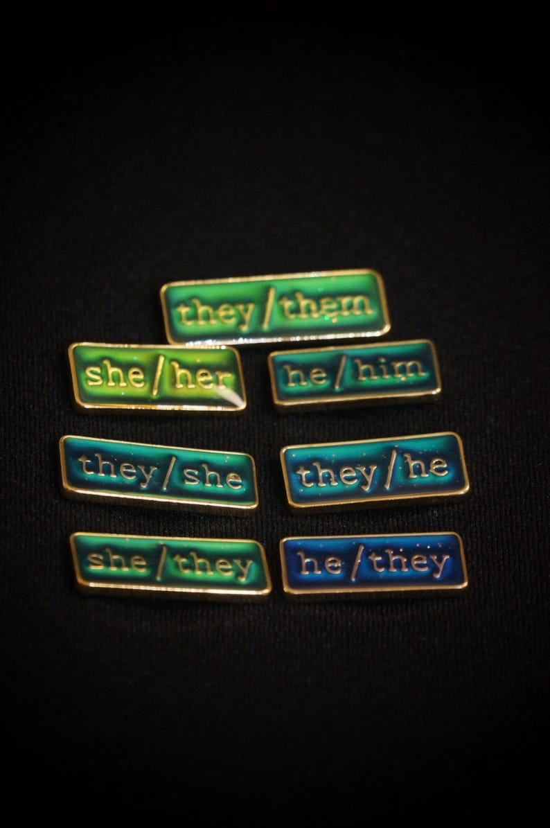 A collection of seven rectangular pronoun pins showing every combination of he, she, and they pronouns in gold text. The background colours are variously blue and greens with small flecks of glitter.