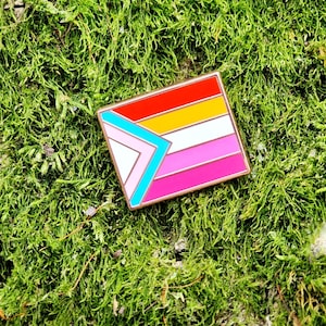 A rectangular hard enamel pin with the trans pride chevron from the progress pride flag on the left side. The rest of the design is the lesbian pride stripes, outlined in rose gold metal.