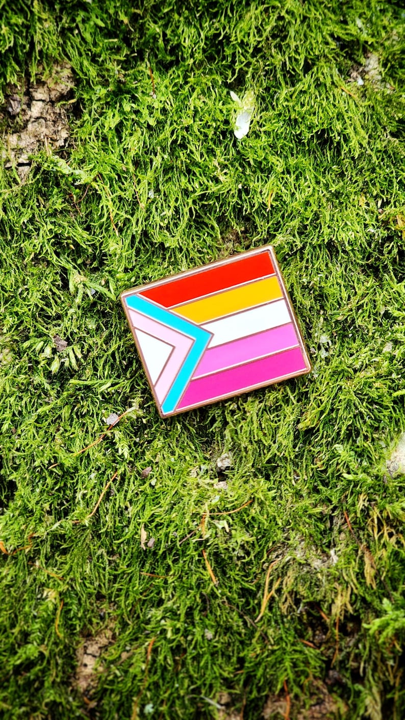 A rectangular hard enamel pin with the trans pride chevron from the progress pride flag on the left side. The rest of the design is the lesbian pride stripes, outlined in rose gold metal.