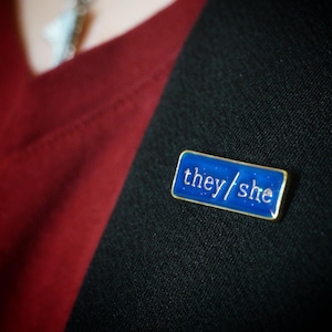 The they/she badge on a lapel. The background looks blue