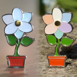 Könnte beinhalten: Zwei Emaille-Anstecknadeln mit einer Blume in einem Topf. Die Blume links ist weiß mit hellblauen Akzenten und die Blume rechts ist weiß mit rosa, lila und hellblauen Akzenten.