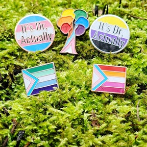 Five enamel pins. Two are the same as in the other photos. Two more are circular and say the words "it's Dr, actually" on either a trans or nonbinary flag background. The final pin is a progress pride flag in the shape of a tree.