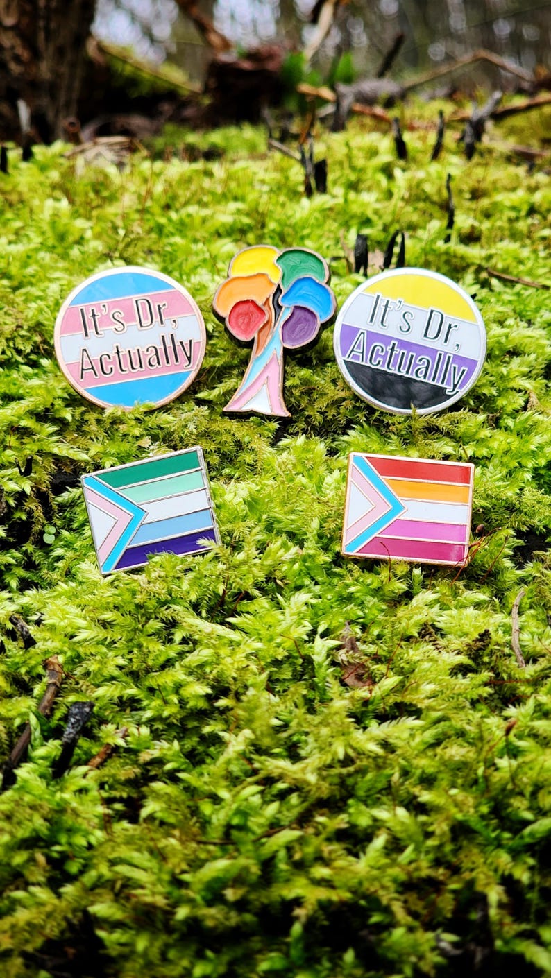 Five enamel pins. Two are the same as in the other photos. Two more are circular and say the words "it's Dr, actually" on either a trans or nonbinary flag background. The final pin is a progress pride flag in the shape of a tree.