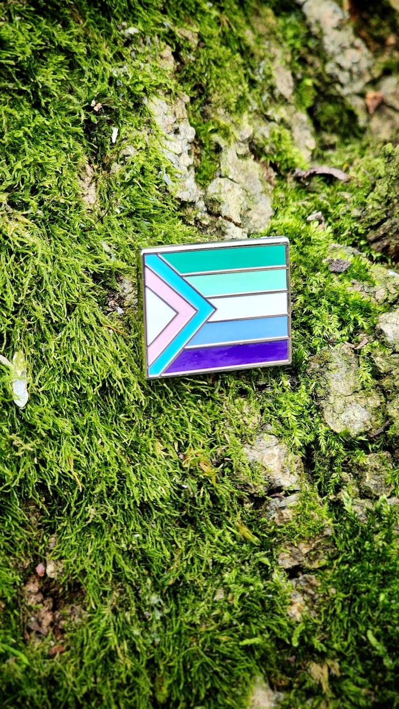 A rectangular hard enamel pin with the trans pride chevron from the progress pride flag on the left side. The rest of the design is the gay man pride stripes, outlined in silver metal.