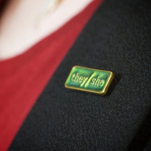 The they/she badge on a lapel. The background looks yellowy green