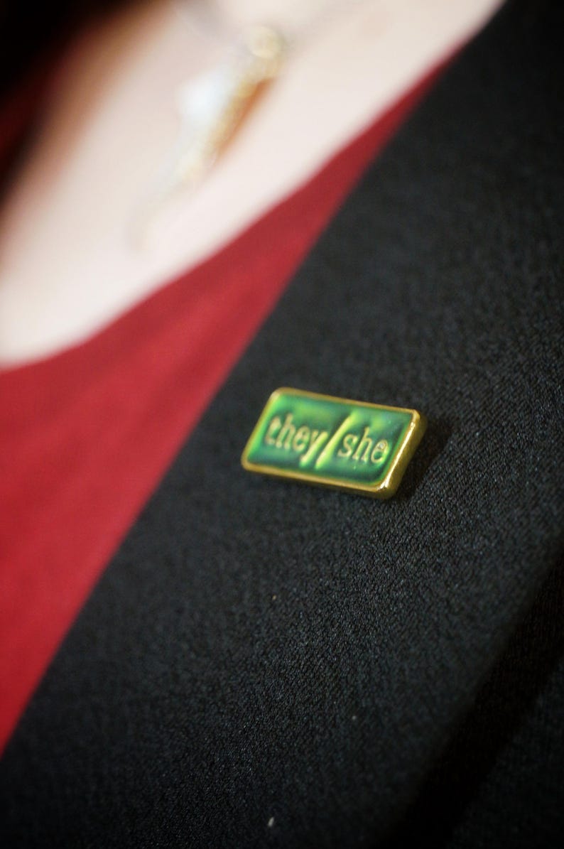 The they/she badge on a lapel. The background looks yellowy green
