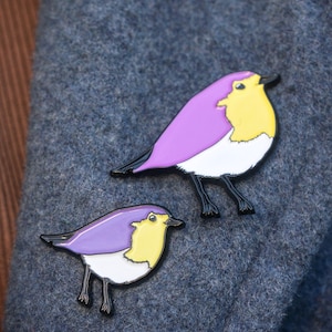 May include: Two bird-shaped enamel pins. The birds have purple wings, yellow chests, white bellies, and black legs and beaks. The pins are set against a textured, dark grey background.