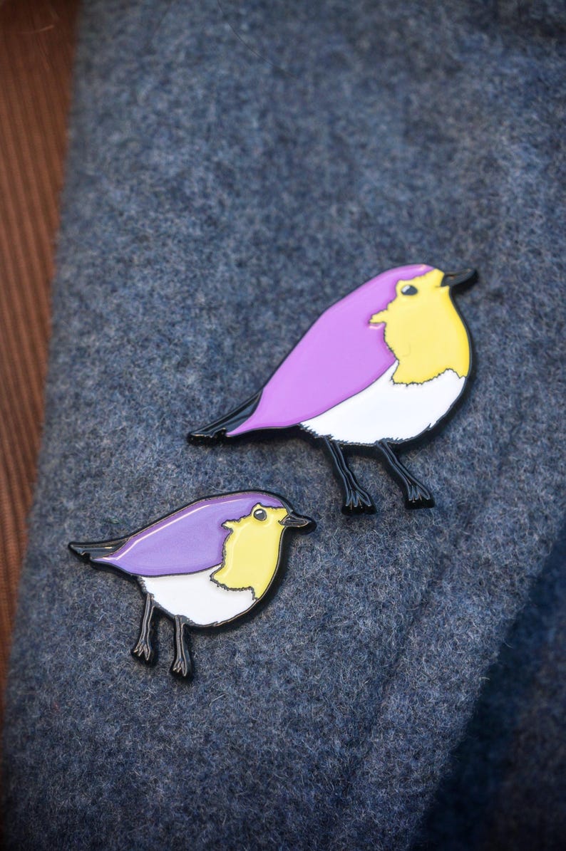 May include: Two bird-shaped enamel pins. The birds have purple wings, yellow chests, white bellies, and black legs and beaks. The pins are set against a textured, dark grey background.