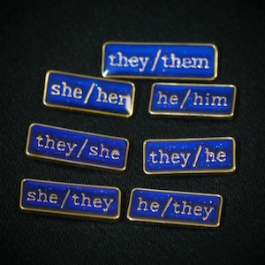A collection of seven rectangular pronoun pins showing every combination of he, she, and they pronouns in gold text. The background colours are deep blue with small flecks of glitter.