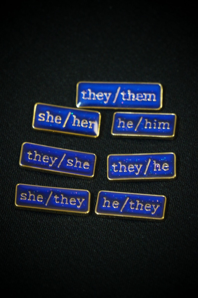 A collection of seven rectangular pronoun pins showing every combination of he, she, and they pronouns in gold text. The background colours are deep blue with small flecks of glitter.