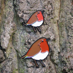 May include: Two enamel robin bird pins. The pins feature a brown back, red breast, white belly, and black details. They are set against a textured tree bark background. The birds are facing left.