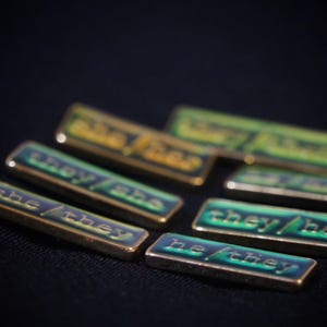 A collection of seven rectangular pronoun pins showing every combination of he, she, and they pronouns in gold text. The background colours are variously blue, greens, yellows, and oranges with small flecks of glitter.