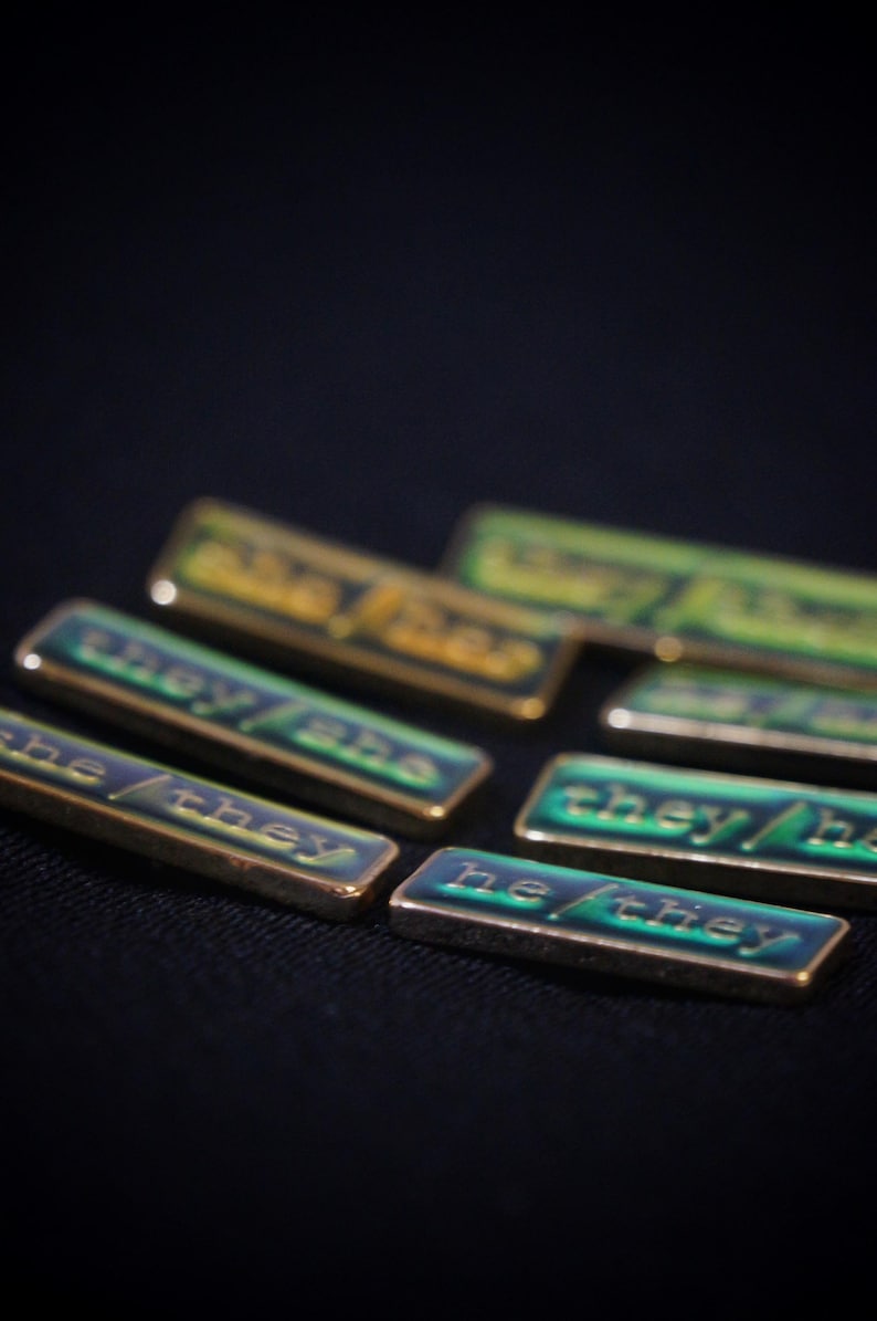A collection of seven rectangular pronoun pins showing every combination of he, she, and they pronouns in gold text. The background colours are variously blue, greens, yellows, and oranges with small flecks of glitter.