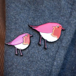 May include: Two pink bird pins with black outlines and legs. The birds have white bellies and orange-red chests. The pins are displayed on a textured, blue-grey fabric.