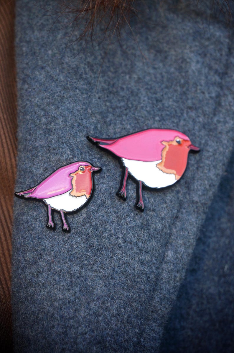May include: Two pink bird pins with black outlines and legs. The birds have white bellies and orange-red chests. The pins are displayed on a textured, blue-grey fabric.