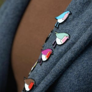 May include: Several bird-shaped enamel pins in shades of blue, green, purple, and brown are pinned to a grey fabric. Each pin has a white outline, creating a charming, decorative effect.