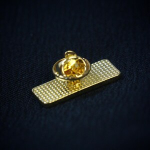May include: A gold-colored pin with a rectangular base and a round, textured top. The pin has a shiny, reflective surface and a small, pointed backing for attachment. The background is a dark blue fabric.