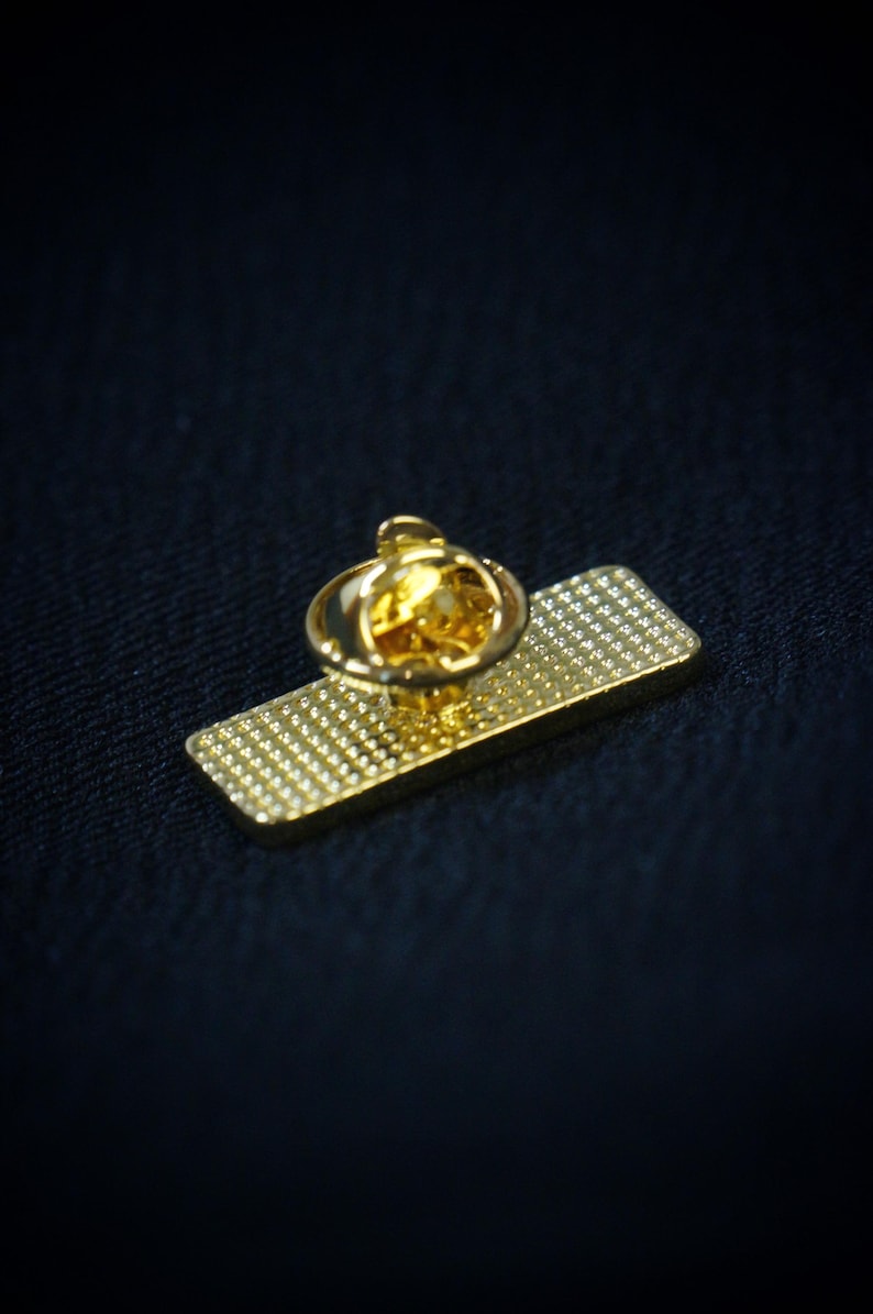 May include: A gold-colored pin with a rectangular base and a round, textured top. The pin has a shiny, reflective surface and a small, pointed backing for attachment. The background is a dark blue fabric.