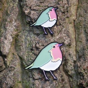May include: Two bird-shaped enamel pins. Each pin features a robin design with a mint green body, a pink breast, and a white belly. The pins have black outlines and legs, set against a textured tree bark background.