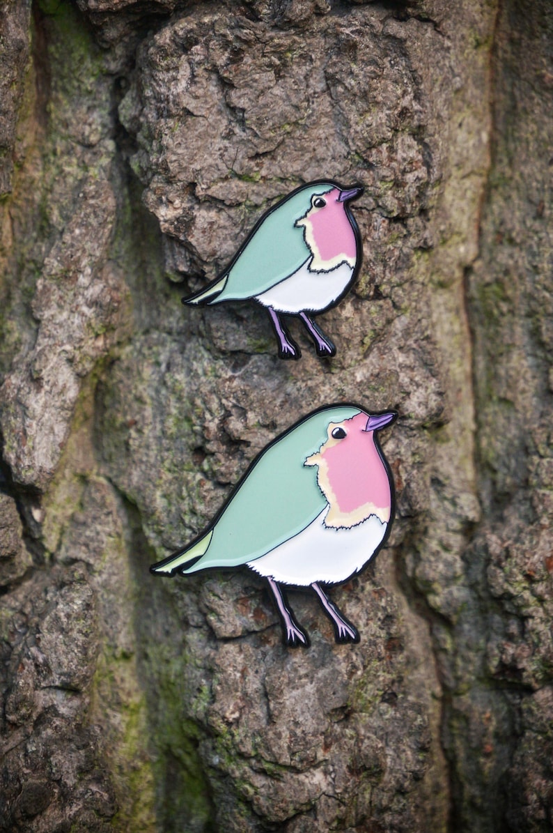 May include: Two bird-shaped enamel pins. Each pin features a robin design with a mint green body, a pink breast, and a white belly. The pins have black outlines and legs, set against a textured tree bark background.
