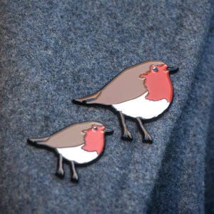May include: Two robin bird enamel pins. The pins have a brown body, white chest, and a red breast. The birds have black legs and beaks. The pins are set against a textured, dark blue-grey background.