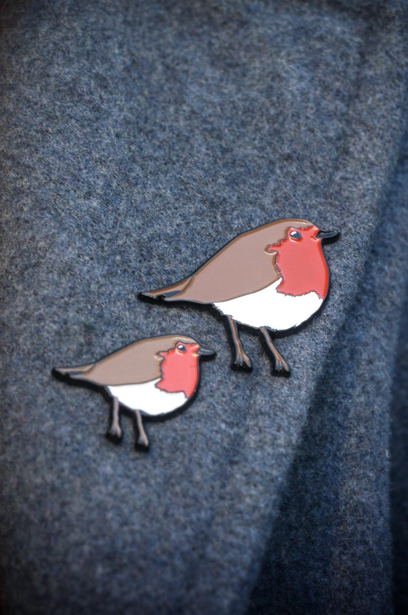 May include: Two robin bird enamel pins. The pins have a brown body, white chest, and a red breast. The birds have black legs and beaks. The pins are set against a textured, dark blue-grey background.
