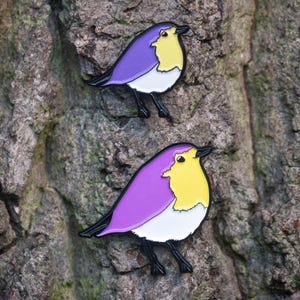 May include: Two bird-shaped enamel pins with a purple back, yellow chest, white belly, and black legs and beak. The pins are attached to a textured, grey tree trunk. The bird pins are a decorative accessory.