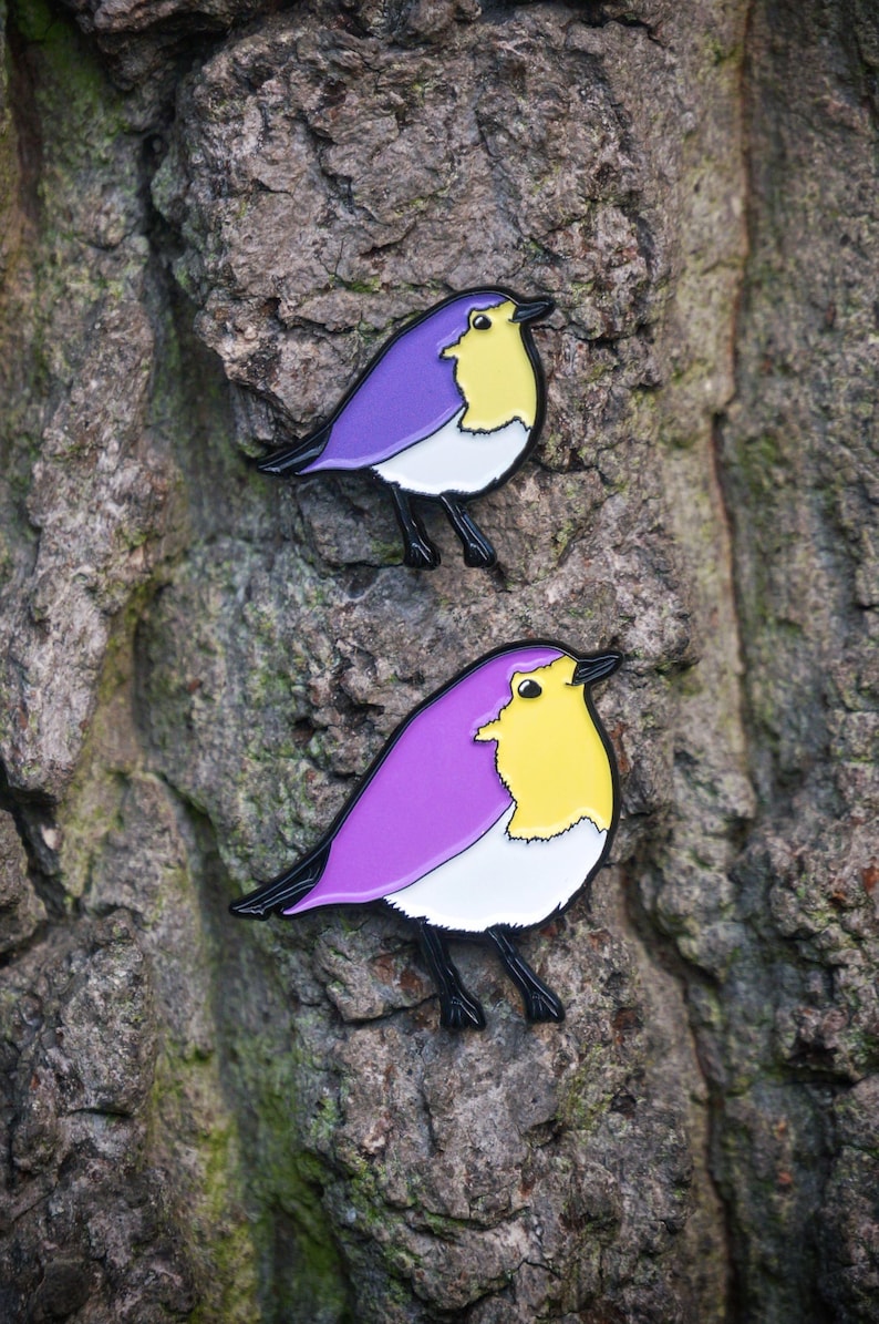 May include: Two bird-shaped enamel pins with a purple back, yellow chest, white belly, and black legs and beak. The pins are attached to a textured, grey tree trunk. The bird pins are a decorative accessory.