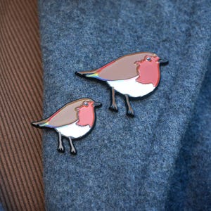 May include: Two robin bird enamel pins. The birds have brown wings, red breasts, white bellies, and black legs. The pins are displayed on a blue wool coat.