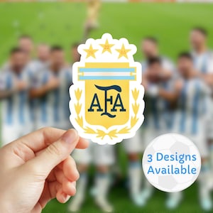 Puede incluir: Una pegatina con el escudo de la Asociación del Fútbol Argentino (AFA), en amarillo y azul. El escudo incluye las letras "AFA" y tres estrellas. También hay un gráfico de un balón de fútbol con el texto "3 Designs Available".