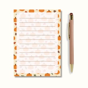 May include: A rectangular notepad featuring a repeating pattern of orange pumpkins and brown leaves on a cream background with horizontal lines for writing, accompanied by a slim, light pink retractable ballpoint pen with gold accents.
