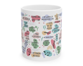 Pode incluir: Caneca de cerâmica branca com afirmações coloridas e frases inspiradoras. A caneca apresenta frases como "Eu sou suficiente", "Eu sou forte" e "Eu mereço ocupar espaço". O design inclui elementos florais e geométricos.