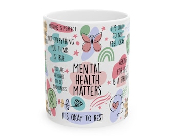 Pode incluir: Caneca de cerâmica branca com as palavras "Mental Health Matters" em um design colorido. A caneca apresenta várias frases e ilustrações relacionadas à saúde mental, incluindo "It's okay to rest" e "Asking for help is a strength."