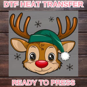 May include: A cartoon reindeer face with brown antlers, a red nose, and a green hat. The text "DTF HEAT TRANSFER" and "READY TO PRESS" are displayed above and below the reindeer image, respectively. Snowflakes are scattered around the reindeer.