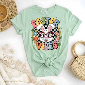 Może przedstawiać: Jasnozielony t-shirt z grafiką przedstawiającą królika w okularach przeciwsłonecznych na tle kratki. Tekst "Easter Vibes" jest napisany w kolorach tęczy.