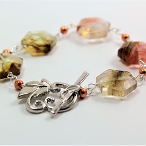 Pode incluir: Uma pulseira de pedras preciosas com pedras facetadas multicoloridas em tons de castanho, rosa e amarelo. As pedras s&atilde;o conectadas por uma corrente prateada e pequenas contas cor de cobre. A pulseira tem um fecho decorativo prateado.