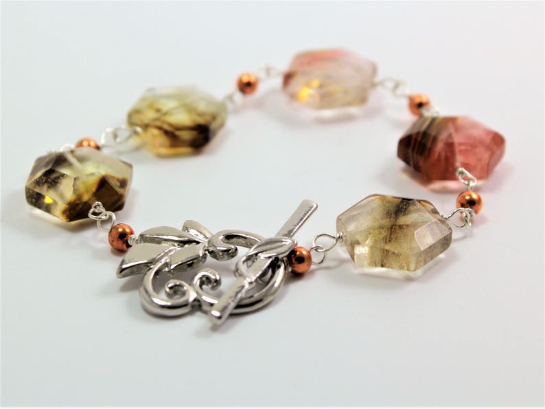 Pode incluir: Uma pulseira de pedras preciosas com pedras facetadas multicoloridas em tons de castanho, rosa e amarelo. As pedras s&atilde;o conectadas por uma corrente prateada e pequenas contas cor de cobre. A pulseira tem um fecho decorativo prateado.