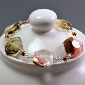 Pode incluir: Uma pulseira de pedras preciosas com pedras facetadas multicoloridas, incluindo tons rosa, castanho e amarelo-esverdeado, enfiadas numa corrente prateada com contas cor de cobre. A pulseira est&aacute; sobre uma chaleira branca.