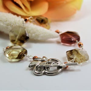 Pode incluir: Uma pulseira de pedras preciosas com contas facetadas de formato irregular em tons de castanho, &acirc;mbar e rosa. A pulseira &eacute; enfiada em uma corrente prateada com detalhes em cobre e um fecho prateado decorativo. Um objeto semelhante a coral e uma flor ao fundo.