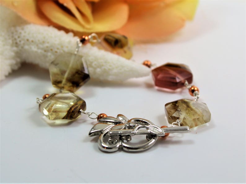 Pode incluir: Uma pulseira de pedras preciosas com contas facetadas de formato irregular em tons de castanho, &acirc;mbar e rosa. A pulseira &eacute; enfiada em uma corrente prateada com detalhes em cobre e um fecho prateado decorativo. Um objeto semelhante a coral e uma flor ao fundo.