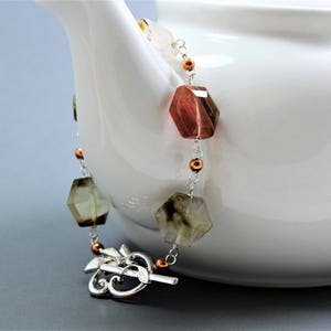 Pode incluir: Um colar de pedras preciosas com contas geom&eacute;tricas facetadas em tons de castanho, &acirc;mbar e transparente. As contas s&atilde;o enfiadas numa corrente prateada com detalhes em cobre. O colar est&aacute; drapeado sobre o lado de uma chaleira branca.