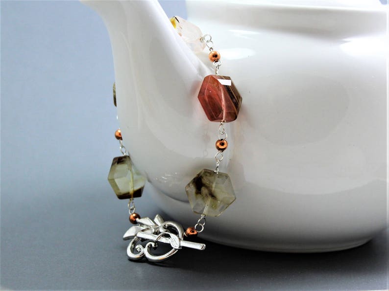 Pode incluir: Um colar de pedras preciosas com contas geom&eacute;tricas facetadas em tons de castanho, &acirc;mbar e transparente. As contas s&atilde;o enfiadas numa corrente prateada com detalhes em cobre. O colar est&aacute; drapeado sobre o lado de uma chaleira branca.