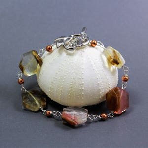 Pode incluir: Uma pulseira artesanal com contas de pedras preciosas multifacetadas e multicoloridas, enfiadas em fio prateado. A pulseira tem pequenas contas cor de cobre e um fecho decorativo. &Eacute; exibida sobre um objeto branco texturizado sobre um fundo cinza.