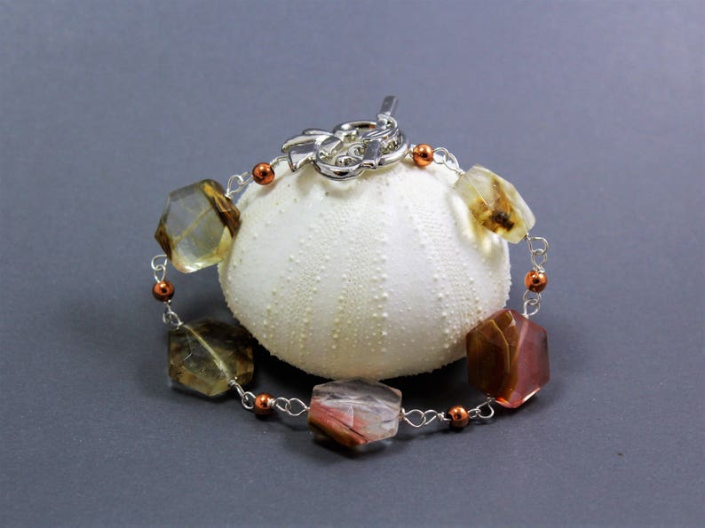 Pode incluir: Uma pulseira artesanal com contas de pedras preciosas multifacetadas e multicoloridas, enfiadas em fio prateado. A pulseira tem pequenas contas cor de cobre e um fecho decorativo. &Eacute; exibida sobre um objeto branco texturizado sobre um fundo cinza.