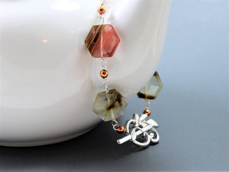 Pode incluir: Infusor de ch&aacute; decorativo com uma corrente de pedras preciosas hexagonais facetadas em tons de rosa, castanho e transparente. O infusor tem um design floral prateado e est&aacute; preso a uma chaleira branca.