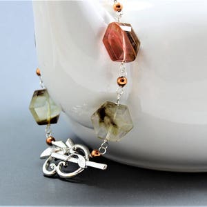 Pode incluir: Um colar de pedras preciosas com contas hexagonais facetadas em tons de castanho, verde e transparente. O colar apresenta uma corrente prateada e um fecho decorativo. Pequenas contas cor de cobre acentuam o design. O colar &eacute; exibido sobre um fundo branco.