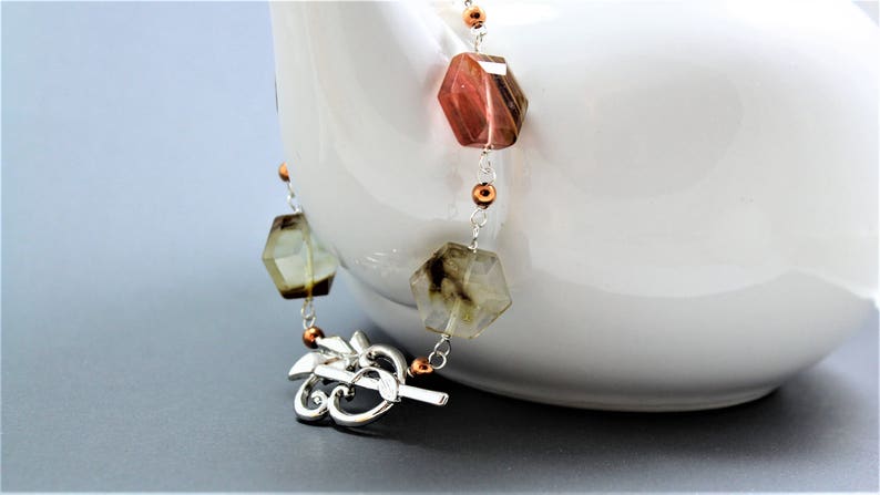 Pode incluir: Um colar de pedras preciosas com contas hexagonais facetadas em tons de castanho, verde e transparente. O colar apresenta uma corrente prateada e um fecho decorativo. Pequenas contas cor de cobre acentuam o design. O colar &eacute; exibido sobre um fundo branco.