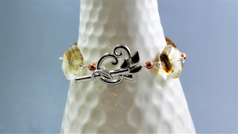 Pode incluir: Uma pulseira de pedras preciosas com fecho prateado. A pulseira apresenta pedras preciosas facetadas, castanho claro e amarelas, conectadas por fio de prata e pequenas contas cor de cobre. O fecho &eacute; um design prateado decorativo.