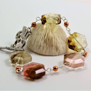 Pode incluir: Uma pulseira de pedras preciosas com pedras multifacetadas multicoloridas e elos de metal prateado. A pulseira apresenta um fecho decorativo e pequenas contas cor de cobre. As pedras s&atilde;o em tons de castanho, rosa e amarelo.