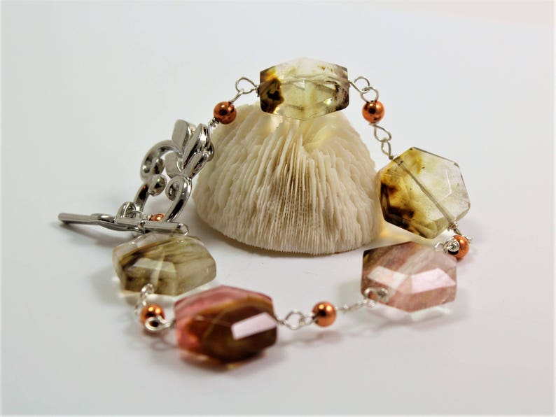 Pode incluir: Uma pulseira de pedras preciosas com pedras multifacetadas multicoloridas e elos de metal prateado. A pulseira apresenta um fecho decorativo e pequenas contas cor de cobre. As pedras s&atilde;o em tons de castanho, rosa e amarelo.